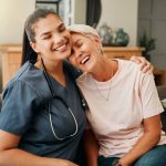 Nurse and elderly woman smiling and embracing on couch, showing care and companionship in a cozy home setting.