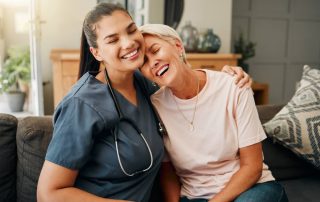 Nurse and elderly woman smiling and embracing on couch, showing care and companionship in a cozy home setting.