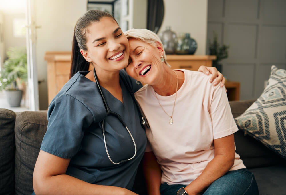 Nurse and elderly woman smiling and embracing on couch, showing care and companionship in a cozy home setting.