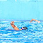 Swimmer in black cap doing freestyle stroke in a blue pool, surrounded by rippling water.