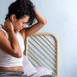 Person sitting on bed feeling neck pain, holding neck with both hands, wearing a white tank top.