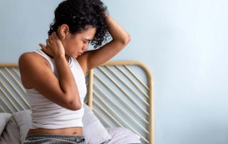 Person sitting on bed feeling neck pain, holding neck with both hands, wearing a white tank top.