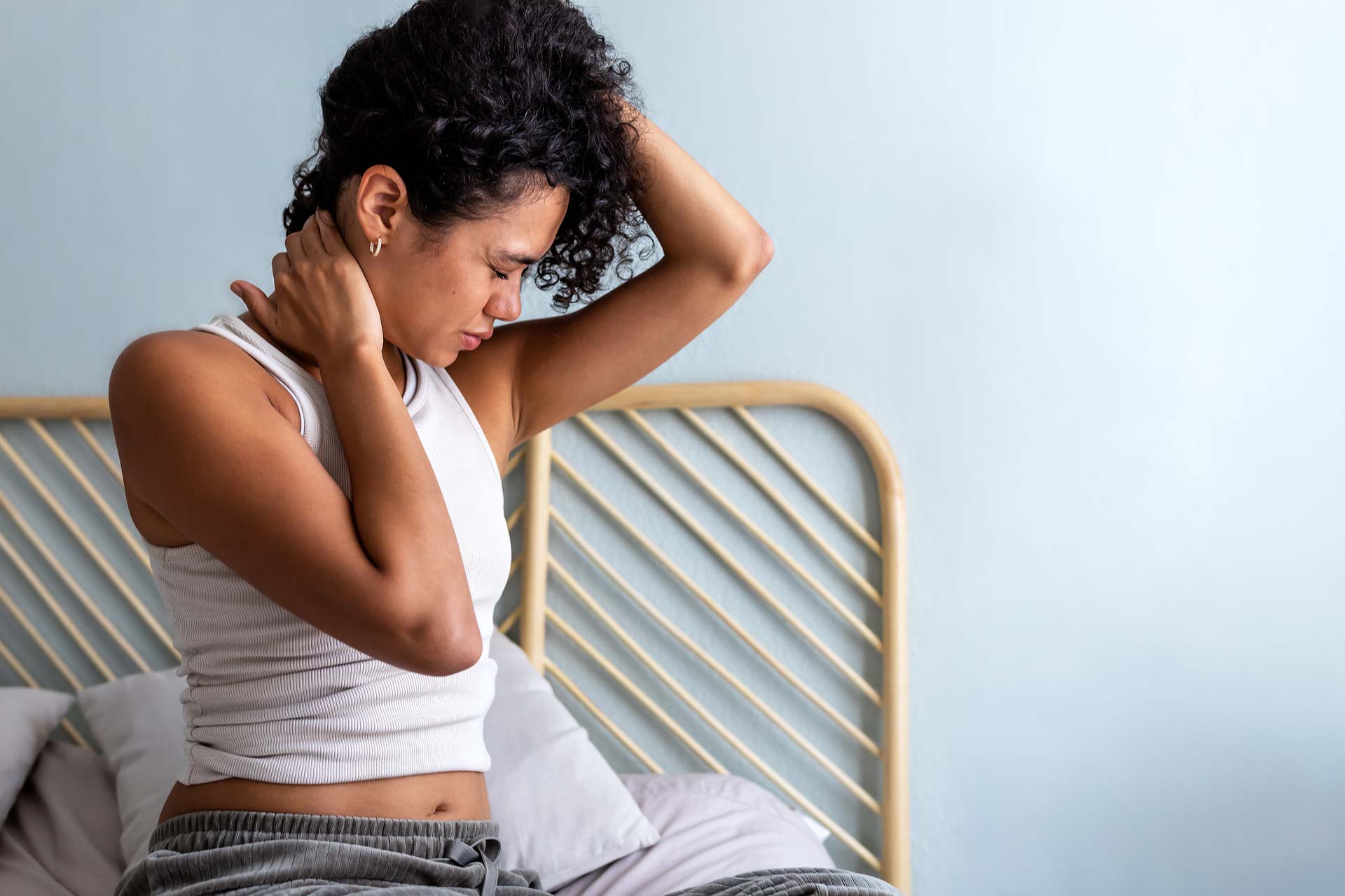 Person sitting on bed feeling neck pain, holding neck with both hands, wearing a white tank top.