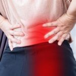 A back view close-up of a person gripping their lower back with both hands, with a prominent red overlay indicating pain on the left side, illustrating symptoms of pelvic floor dysfunction causing back pain.