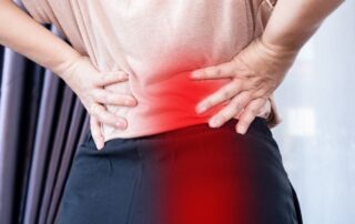 A back view close-up of a person gripping their lower back with both hands, with a prominent red overlay indicating pain on the left side, illustrating symptoms of pelvic floor dysfunction causing back pain.