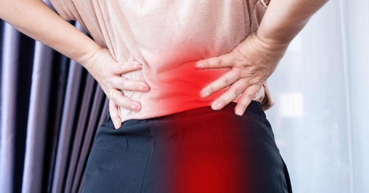 A back view close-up of a person gripping their lower back with both hands, with a prominent red overlay indicating pain on the left side, illustrating symptoms of pelvic floor dysfunction causing back pain.
