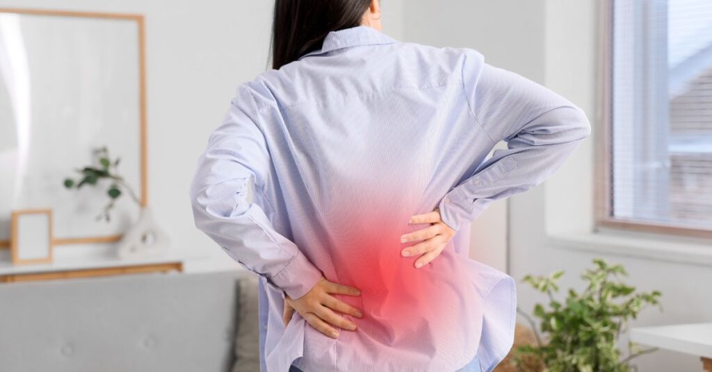 A woman in an office setting sitting at a desk, leaning forward with one hand on her forehead and the other on her lower back in discomfort.